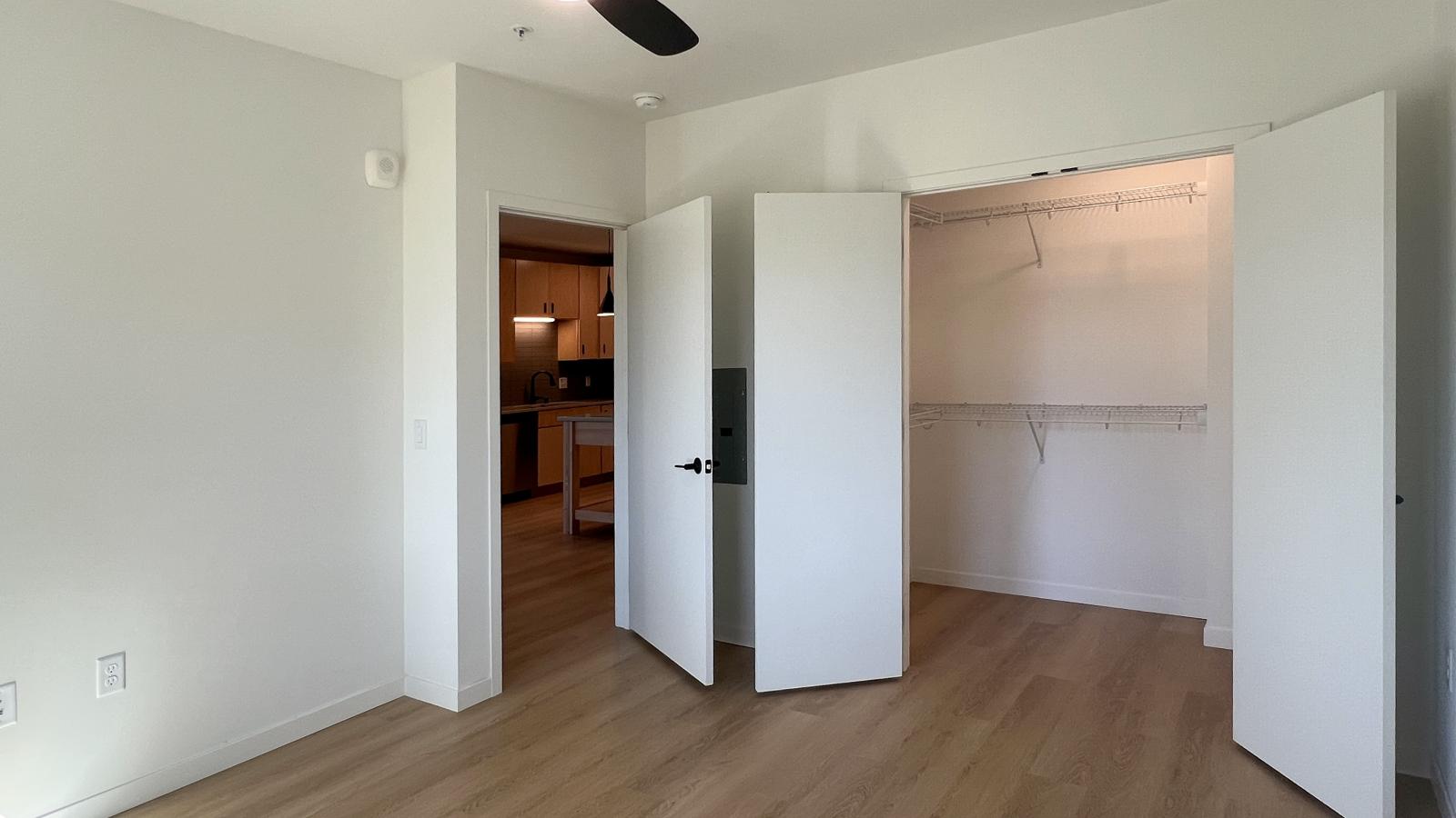 Bedroom in a one bedroom plus den apartment at The Irwin featuring wood-look flooring, ceiling fan, large walk-in closet with built-in shelving, and views toward the kitchen and entry hallway.
