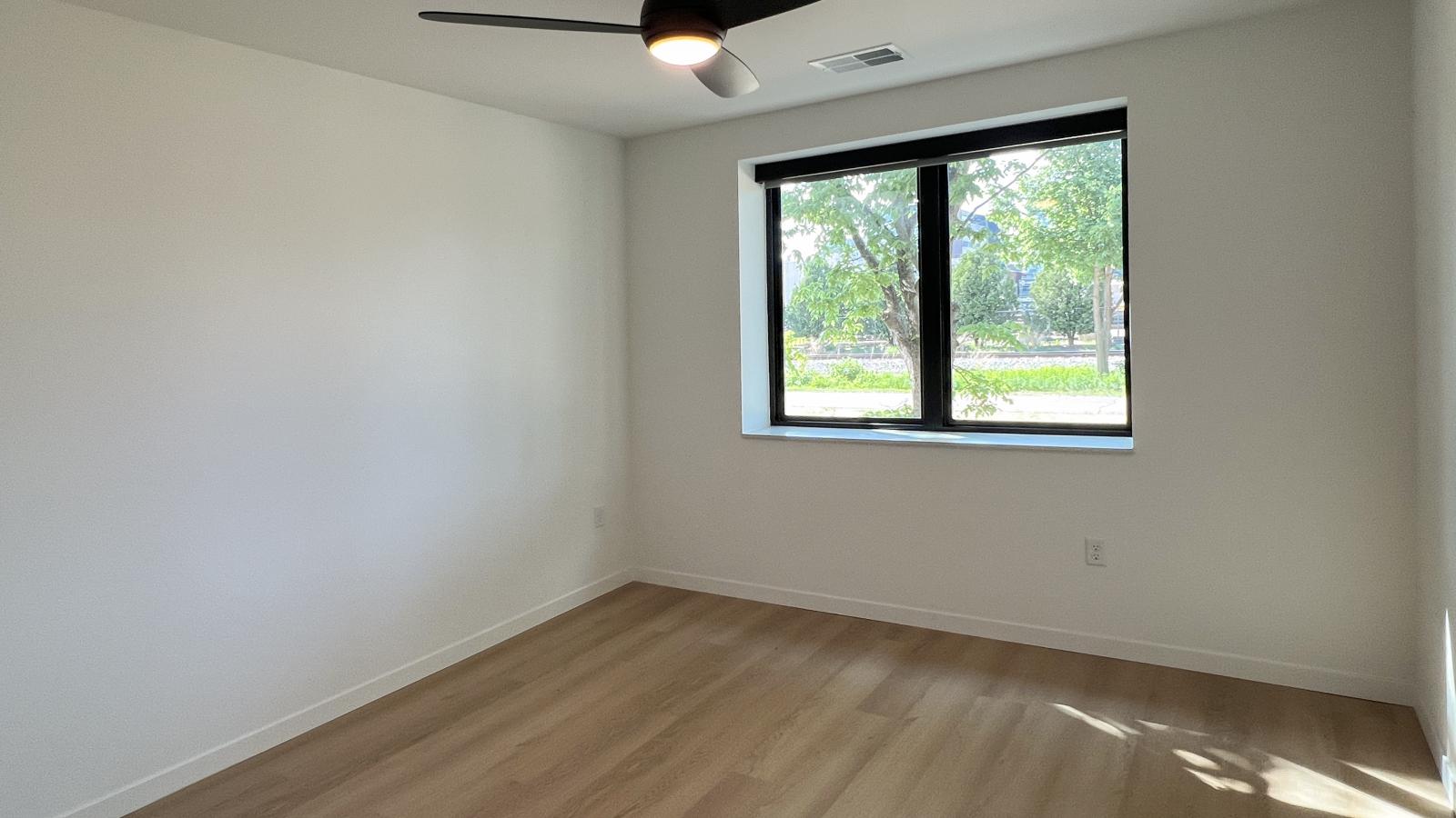Modern one-bedroom plus den apartment bedroom at The Irwin featuring oversized windows, ceiling fan, luxury vinyl flooring, and contemporary finishes in Madison’s Willy Street neighborhood.