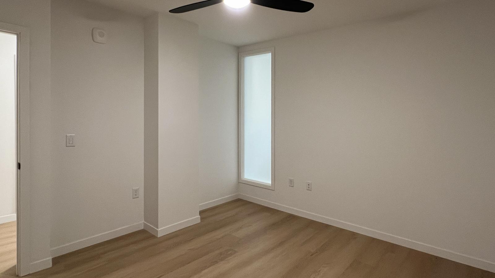 Modern bedroom at The Irwin featuring frosted floor-to-ceiling window, black ceiling fan, luxury vinyl flooring, and clean contemporary design in Madison’s Marquette neighborhood.