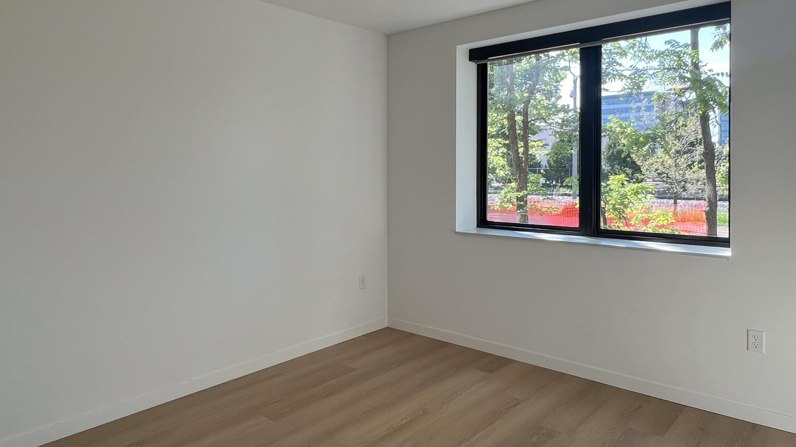 Bright bedroom in a one bedroom plus den apartment at The Irwin with oversized black-trimmed window, ceiling fan, natural light, and modern wood-look flooring.