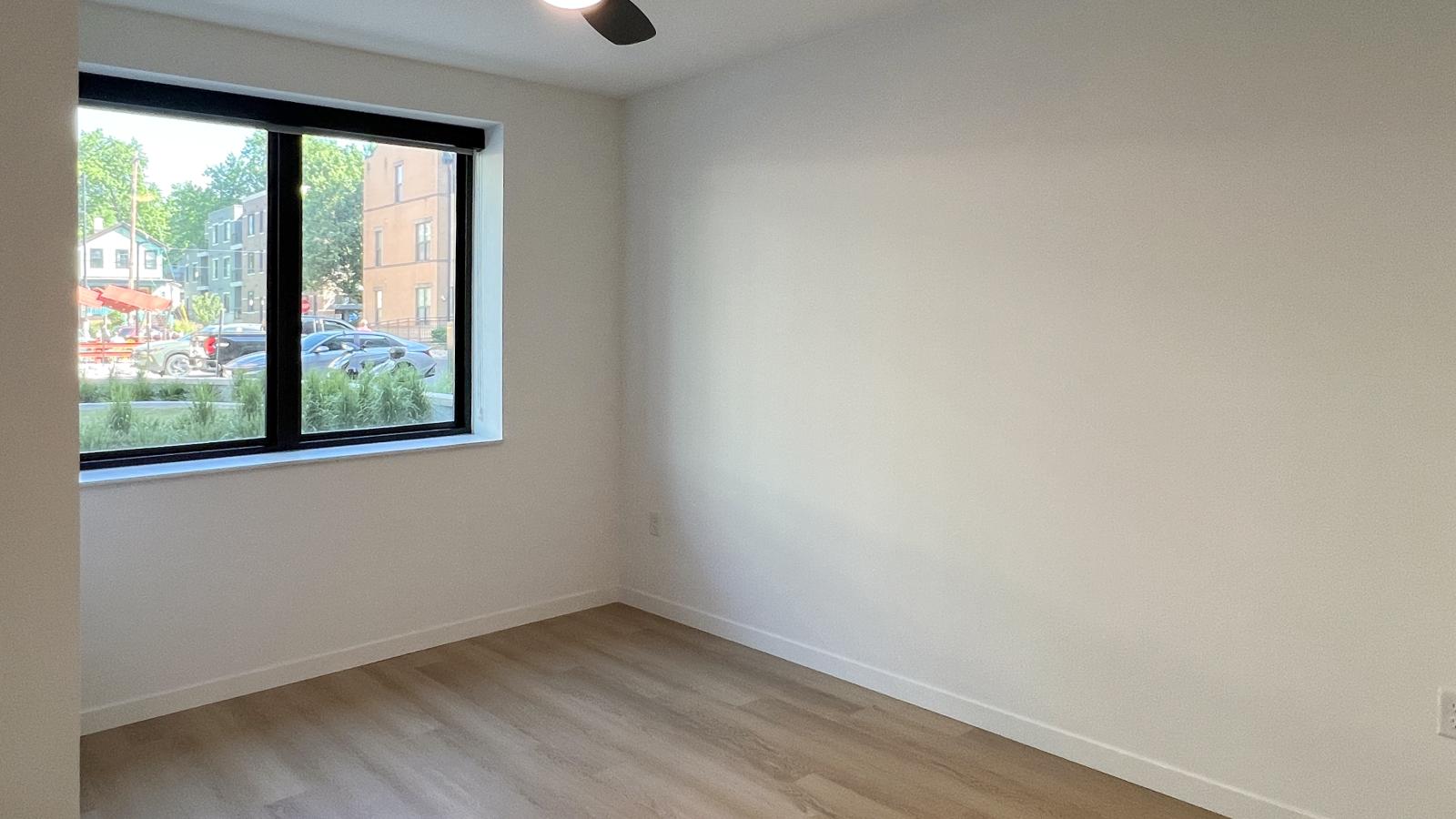 One bedroom + den apartment bedroom with oversized black-framed window, luxury vinyl plank flooring, ceiling fan, and abundant natural light overlooking the neighborhood streetscape in downtown Madison