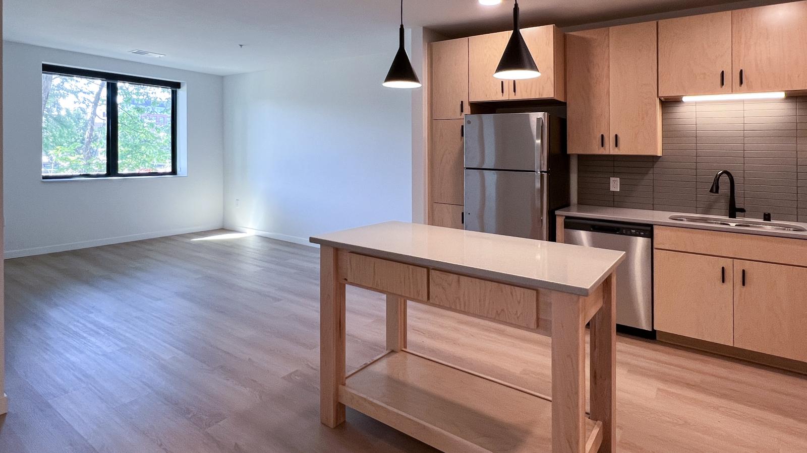 Open-concept kitchen and living area at The Irwin Apartments with natural wood cabinetry, kitchen island, oversized windows, stainless steel appliances, modern pendant lighting, and spacious layout in Madison, Wisconsin.