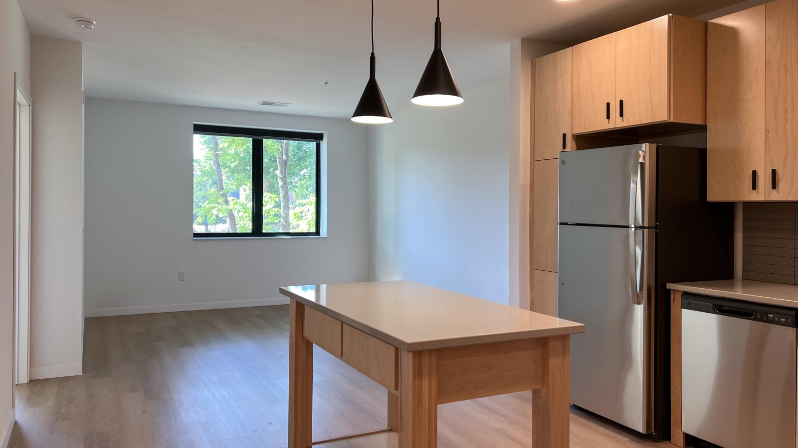 Open concept kitchen and living area in a one bedroom plus den apartment at The Irwin featuring light wood cabinetry, stainless steel appliances, movable island, pendant lighting, and large living room window with tree views.