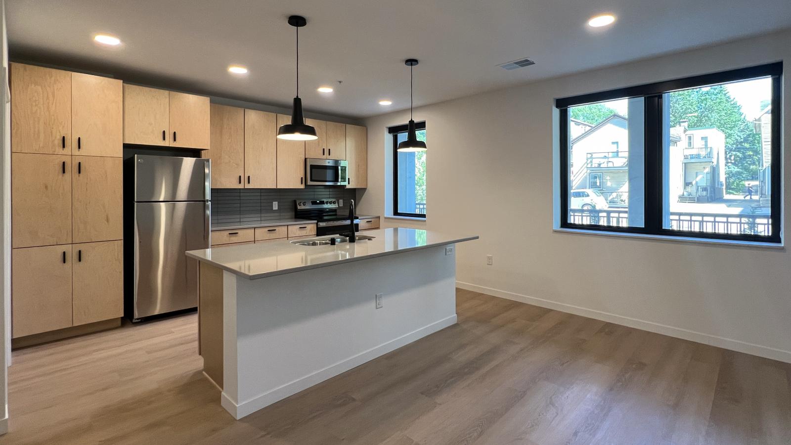 Modern open-concept apartment kitchen and living area at The Irwin featuring quartz countertops, natural wood cabinetry, stainless steel appliances, matte black fixtures, oversized kitchen island, large windows, and luxury vinyl flooring in Madison’s Willy Street neighborhood.