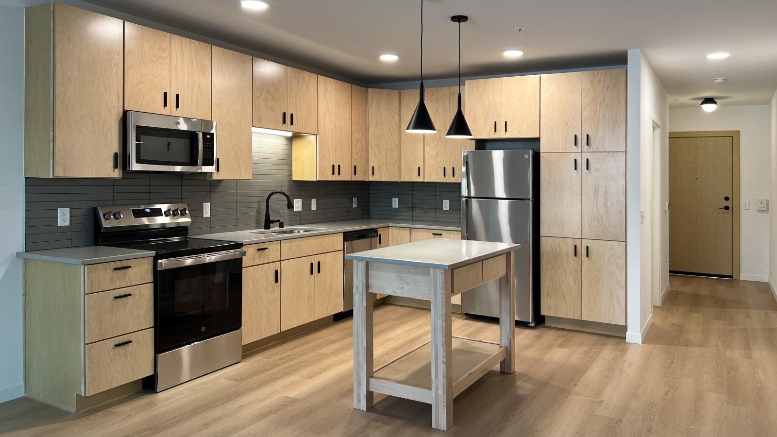 Modern kitchen at The Irwin Apartments with natural wood cabinetry, matte black fixtures, stainless steel appliances, kitchen island, quartz countertops, and luxury vinyl flooring in Madison, Wisconsin.