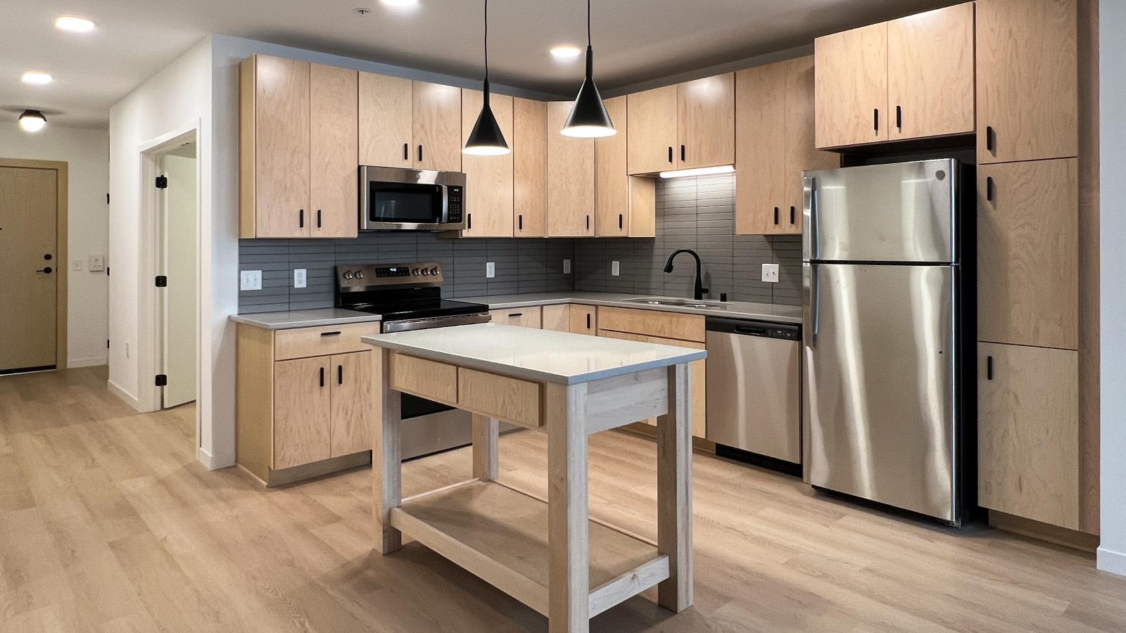 Modern kitchen at The Irwin Apartments featuring natural wood cabinetry, stainless steel appliances, matte black fixtures, kitchen island, quartz countertops, and upscale contemporary finishes in Madison, Wisconsin.