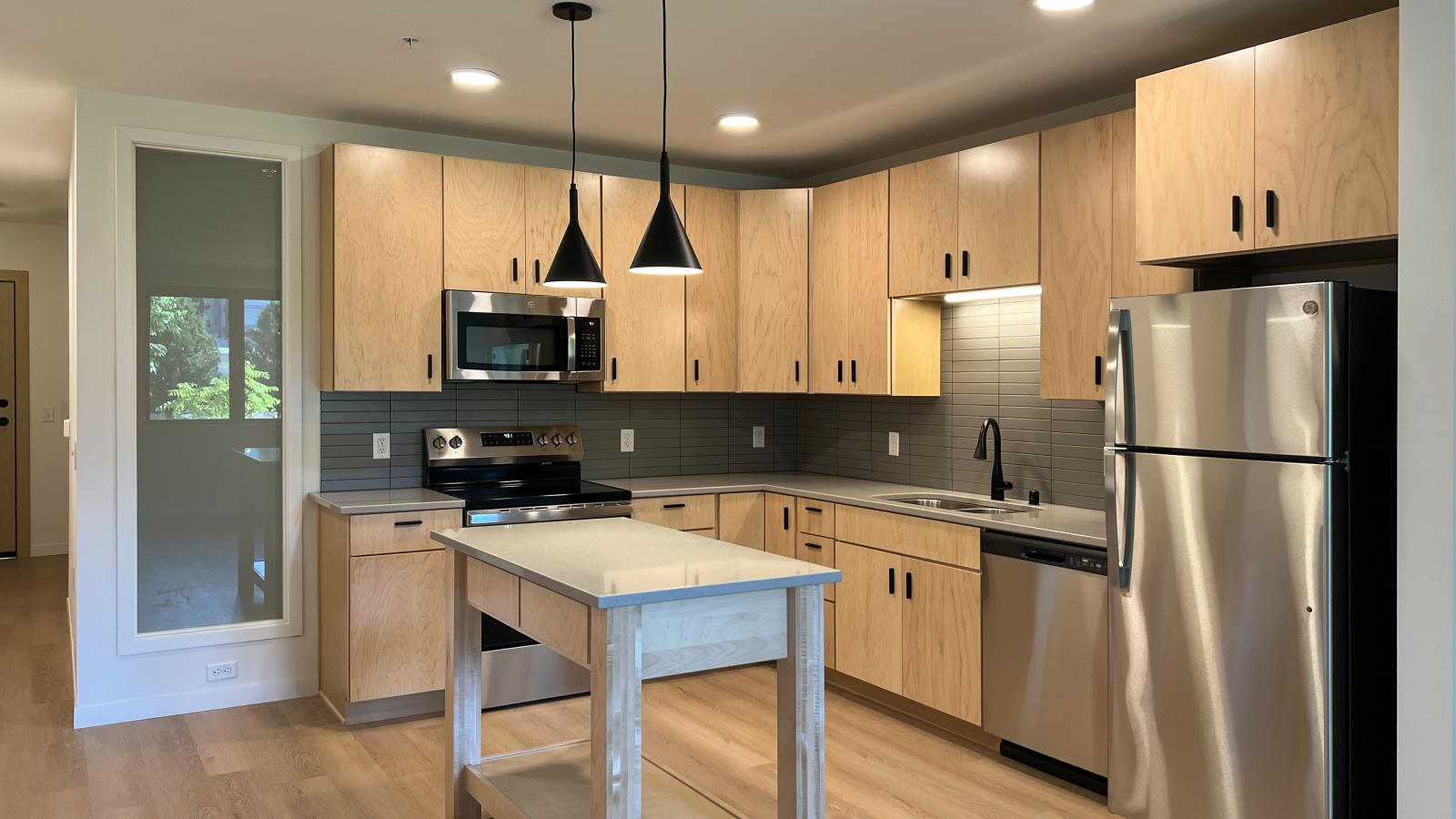 Modern kitchen at The Irwin Apartments featuring natural wood cabinetry, stainless steel appliances, matte black fixtures, quartz countertops, kitchen island, and contemporary finishes in Madison, Wisconsin.