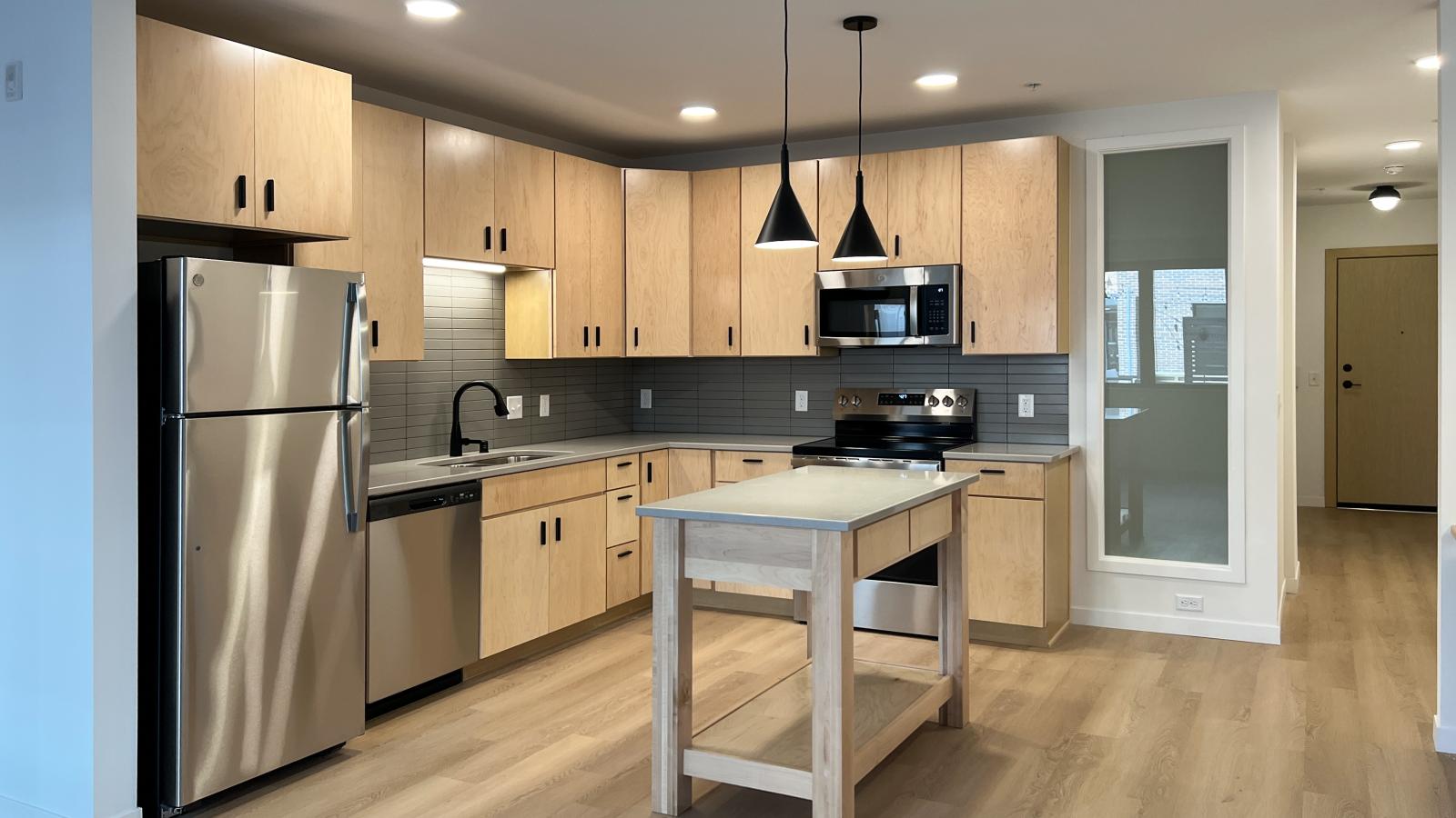 Modern kitchen at The Irwin Apartments with natural wood cabinetry, stainless steel appliances, quartz countertops, matte black fixtures, kitchen island, and upscale contemporary finishes in Madison, Wisconsin.