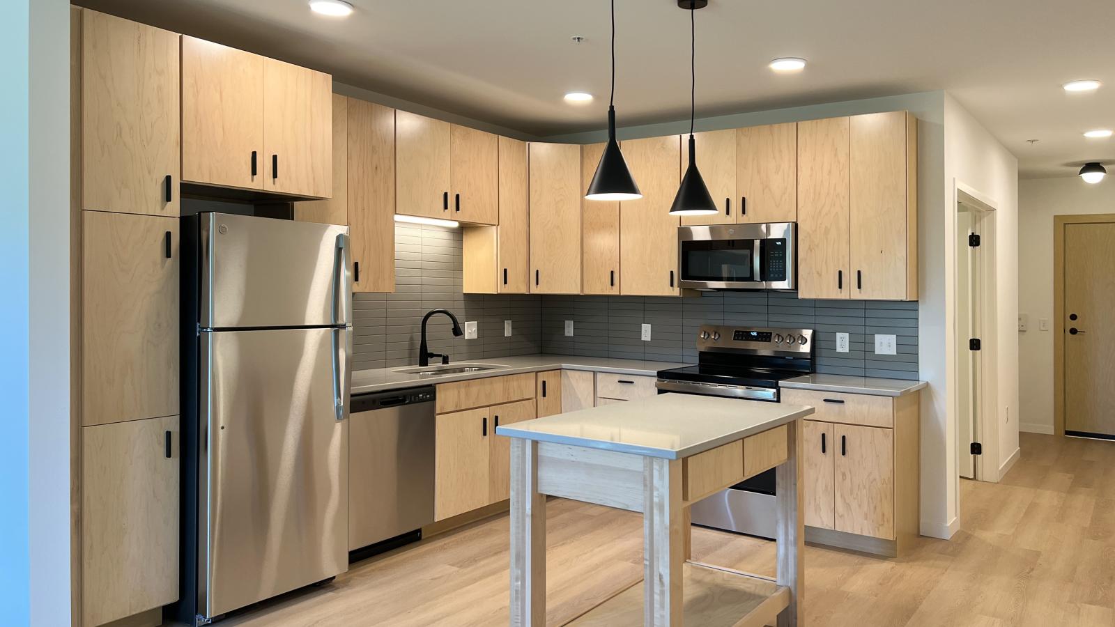 Modern one bedroom plus den apartment kitchen at The Irwin featuring light wood cabinetry, stainless steel appliances, gray tile backsplash, black pendant lighting, and a movable island with open shelving and quartz-style countertops.