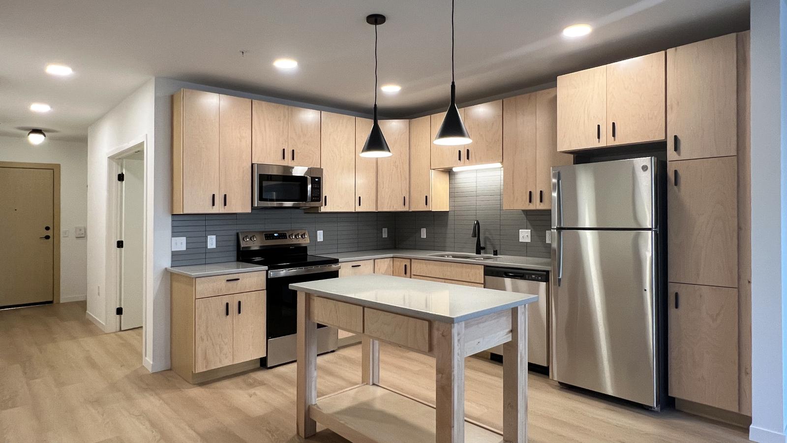 Modern one bedroom + den apartment kitchen with natural wood cabinetry, gray tile backsplash, stainless steel appliances, oversized pantry storage, quartz-style countertops, pendant lighting, and center island in downtown Madison