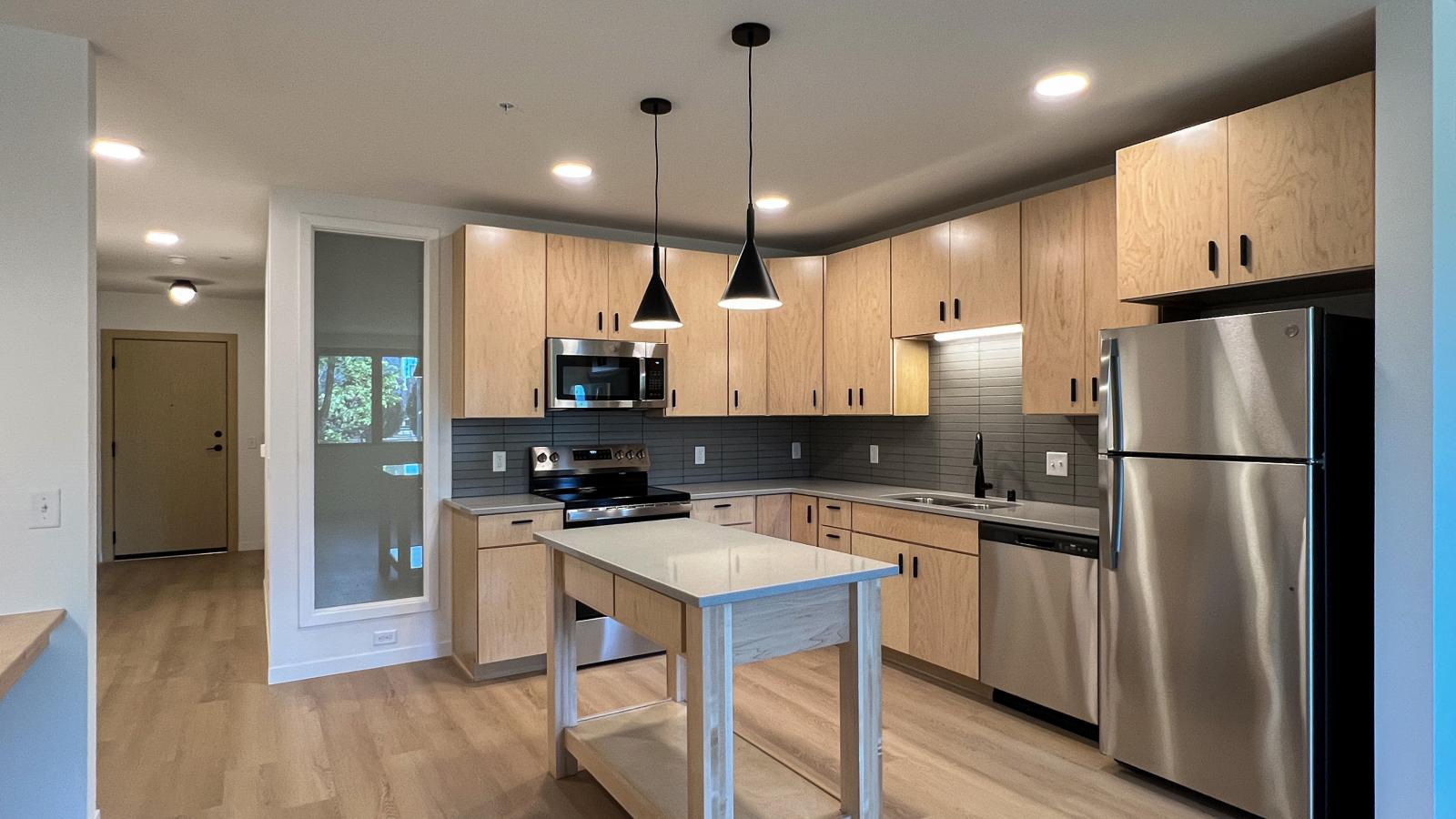 One bedroom apartment featuring an open concept kitchen with natural wood cabinetry, gray tile backsplash, stainless steel appliances, kitchen island, pendant lighting, and built in workspace with modern finishes in downtown Madison