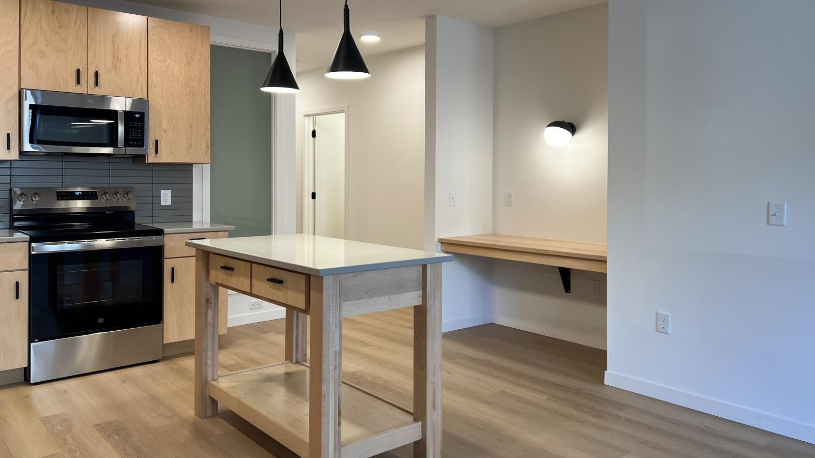 Open-concept kitchen and built-in workspace at The Irwin Apartments featuring natural wood cabinetry, quartz countertops, matte black pendant lighting, stainless steel appliances, and modern finishes in Madison’s Willy Street neighborhood.