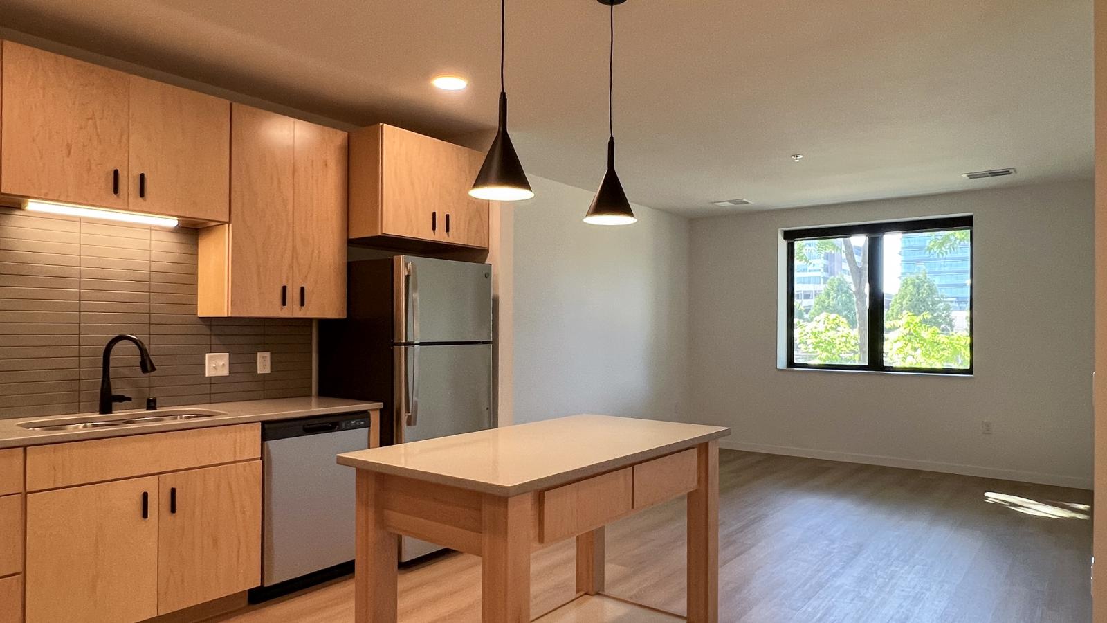 Open-concept living room and kitchen at The Irwin Apartments with oversized window, kitchen island, modern pendant lighting, natural wood cabinetry, and upscale contemporary finishes near downtown Madison.